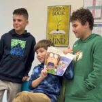 3 students, one holds up a book showing pages to other students.